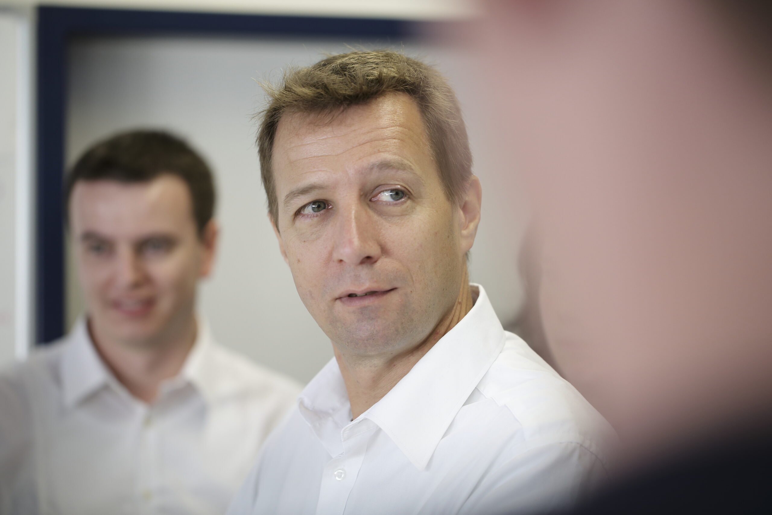 Zwei Männer in weißen Hemden befinden sich in einem Innenraum. Der Mann im Fokus blickt leicht nach rechts, während der andere Mann im Hintergrund lächelt. Das Bild strahlt eine sanfte, professionelle Atmosphäre aus.
