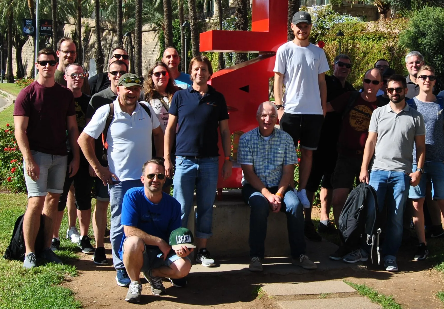 Eine Gruppe von Menschen posiert an einem sonnigen Tag im Freien vor einer großen roten Skulptur, im Hintergrund sind grünes Gras und Palmen zu sehen.