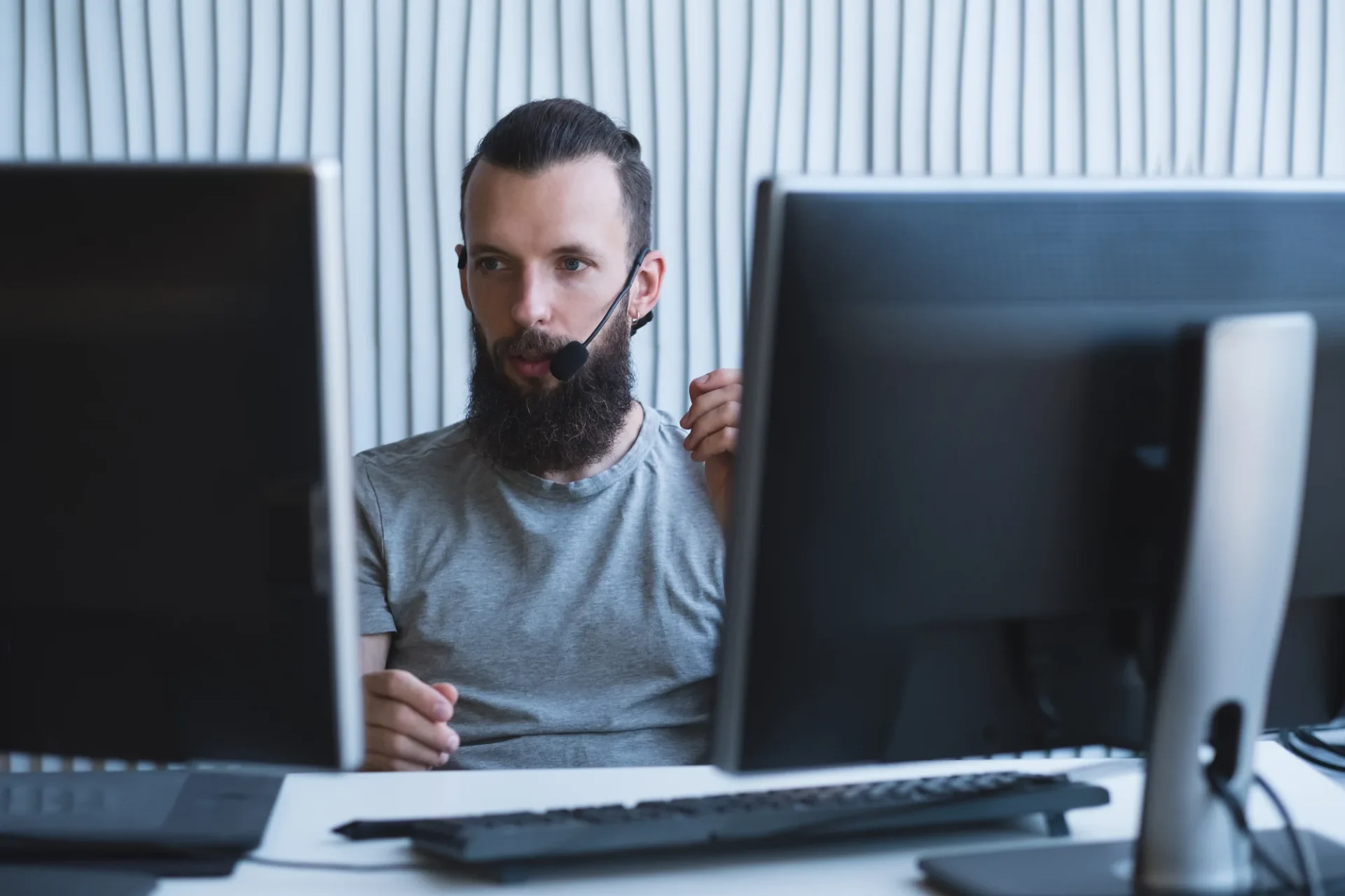 Ein Mann mit Bart und Headset sitzt an einem Schreibtisch zwischen zwei Computermonitoren und wirkt konzentriert, während er arbeitet oder kommuniziert.