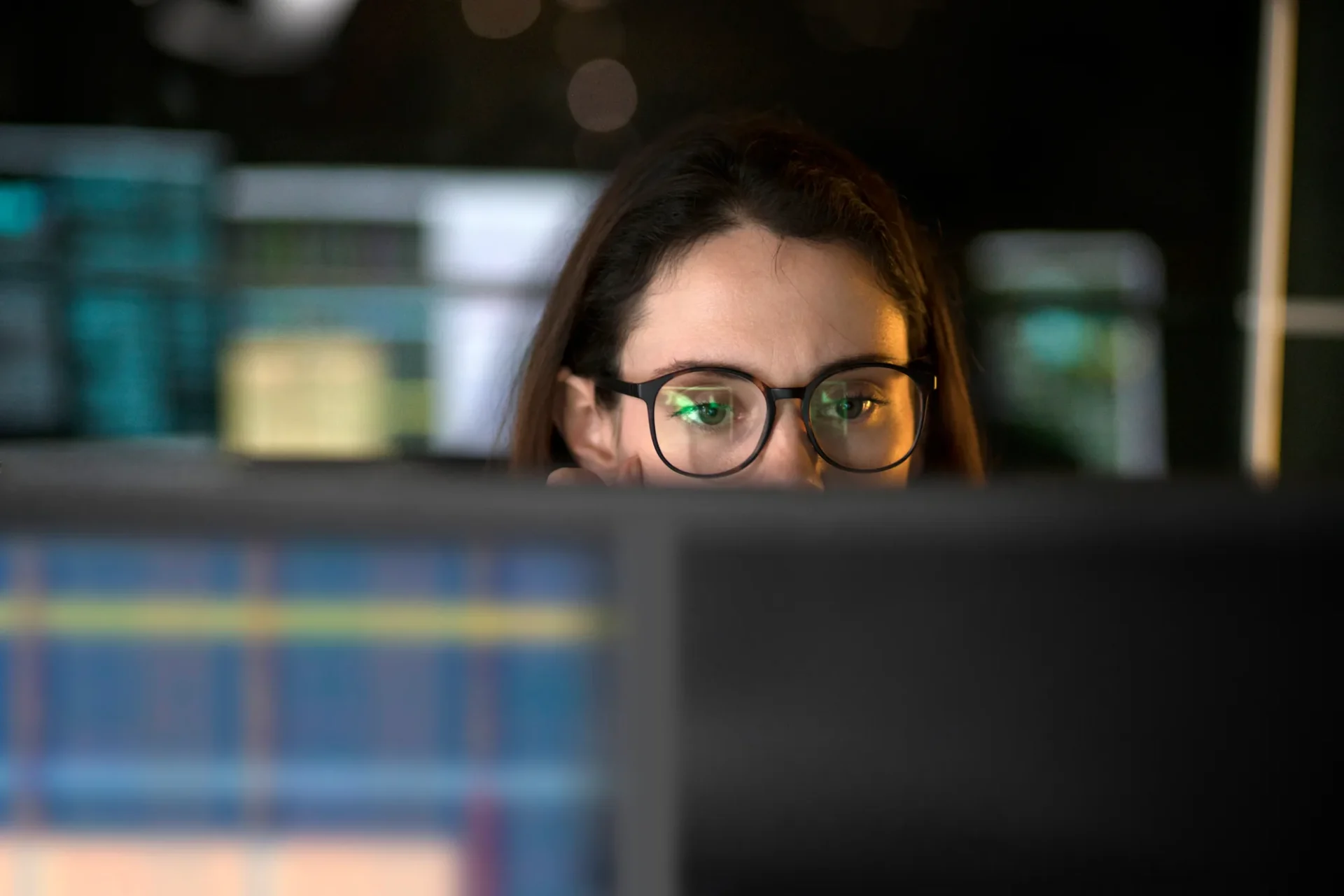 Eine Frau mit Brille betrachtet aufmerksam Computerbildschirme mit Diagrammen oder Daten, was darauf hindeutet, dass sie sich in einer schwach beleuchteten Umgebung auf ihre Arbeit oder eine Analyse konzentriert.