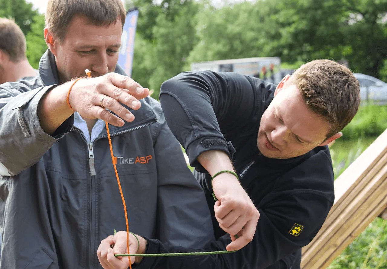 Zwei Männer im Freien sind ganz bei der Sache, während sie sich gegenseitig orangefarbene und grüne Seile um die Handgelenke binden – möglicherweise im Rahmen einer Teambuilding-Maßnahme. Im Hintergrund sind üppiges Grün und verschwommene Teilnehmer zu sehen.